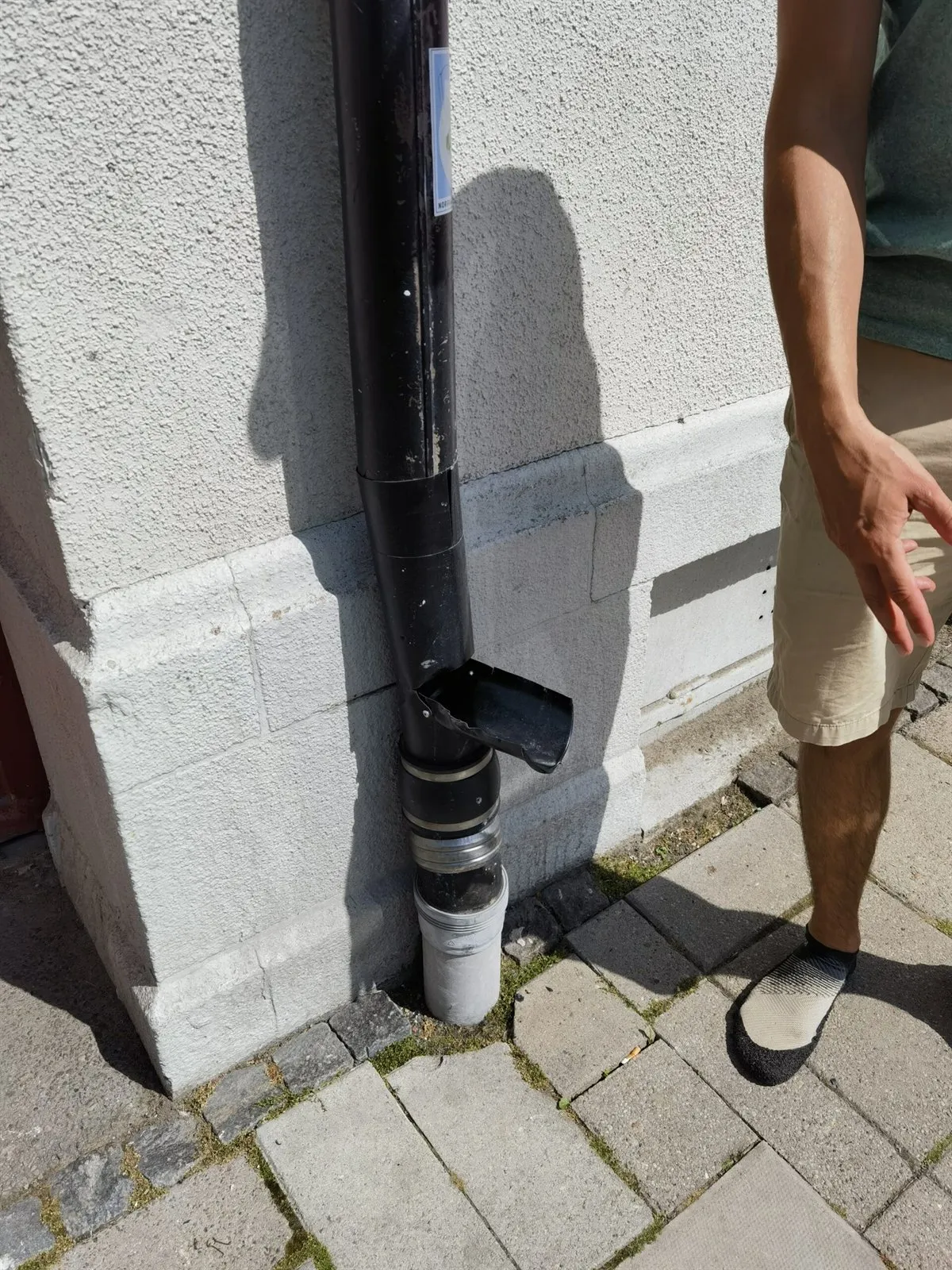 Geöffneter Auslauf an einem Fallrohr neben einer Hausfassade und dem gepflasterten Gehweg