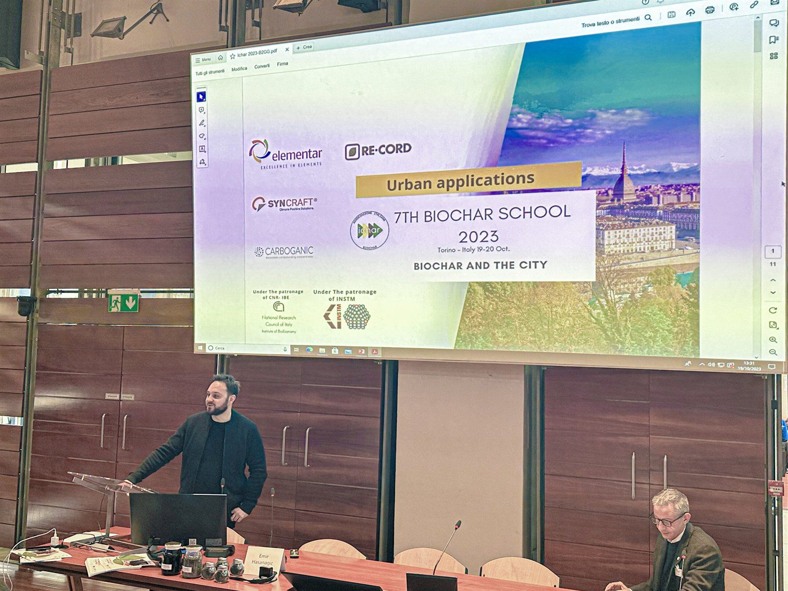 Emir Hasanagić am Vortragsplatz bei der 7th Biochar School 2023, auf der Leinwand die Titelfolie der Veranstaltung